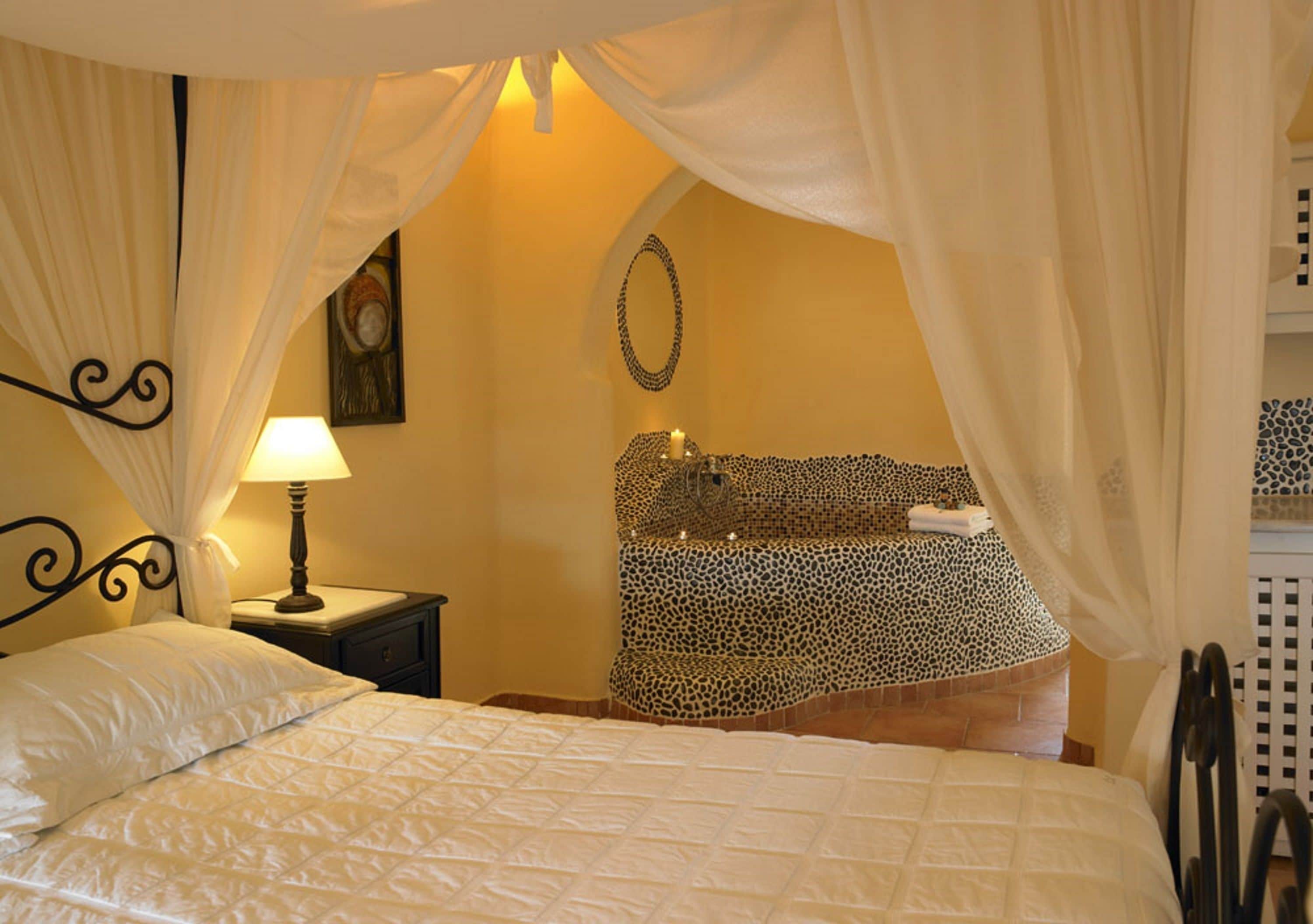 A bedroom with a bed, a nightstand with a lamp, a leopard print bath, and a curtain.