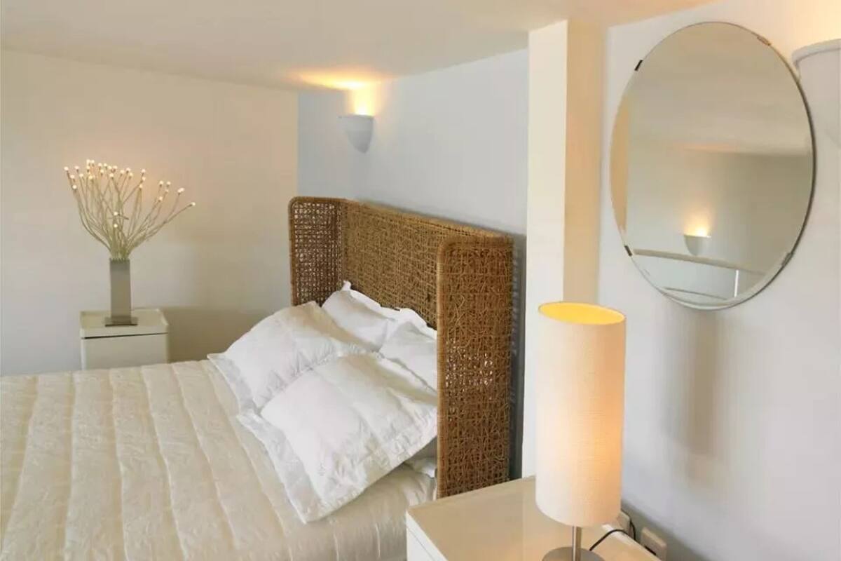 Un lit bien fait, avec du linge de lit blanc, une tête de lit en osier, une table de chevet avec une lampe et un miroir rond accroché au mur.