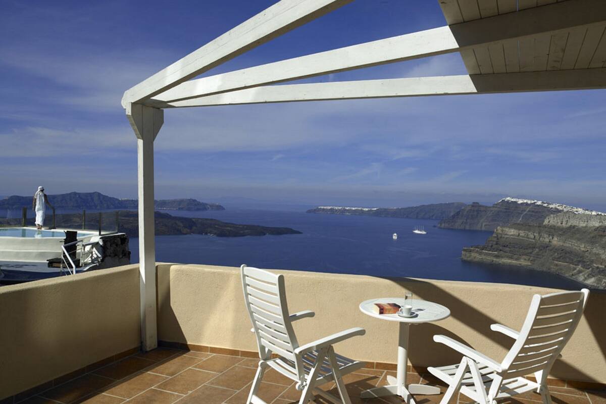 Un balcon avec vue sur la mer et les falaises, des chaises blanches, une petite table et une personne debout près de la piscine.