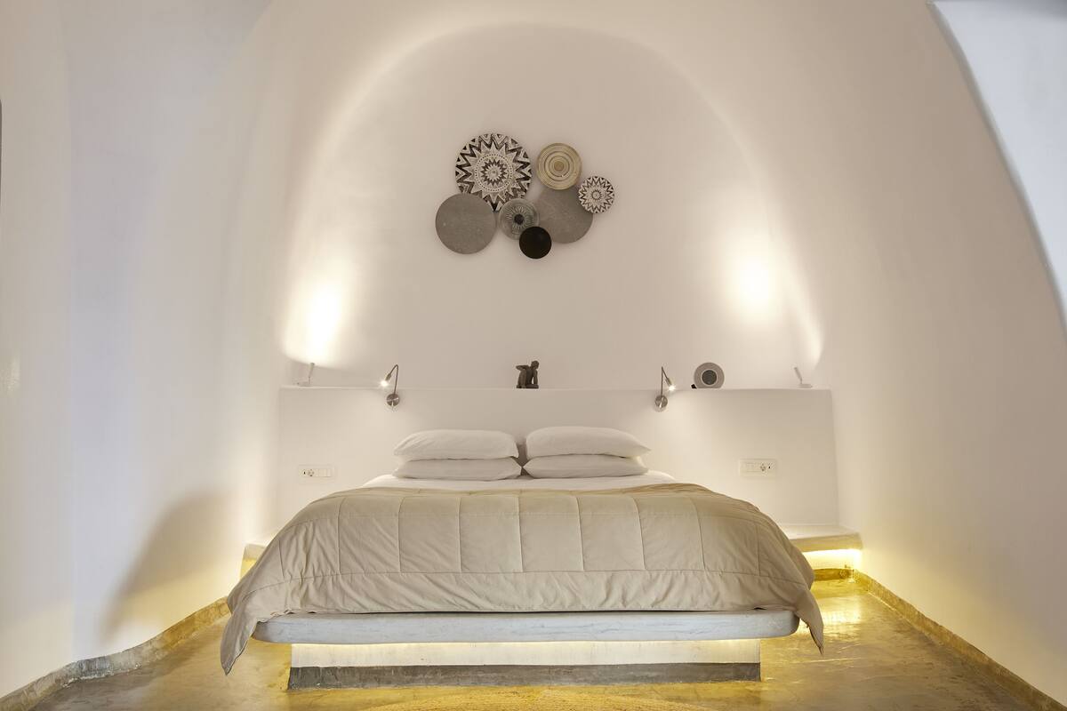 Une chambre à coucher au style minimaliste, avec un lit placé au centre, sous une décoration murale aux motifs géométriques, une table de chevet et une lampe fixée au mur.