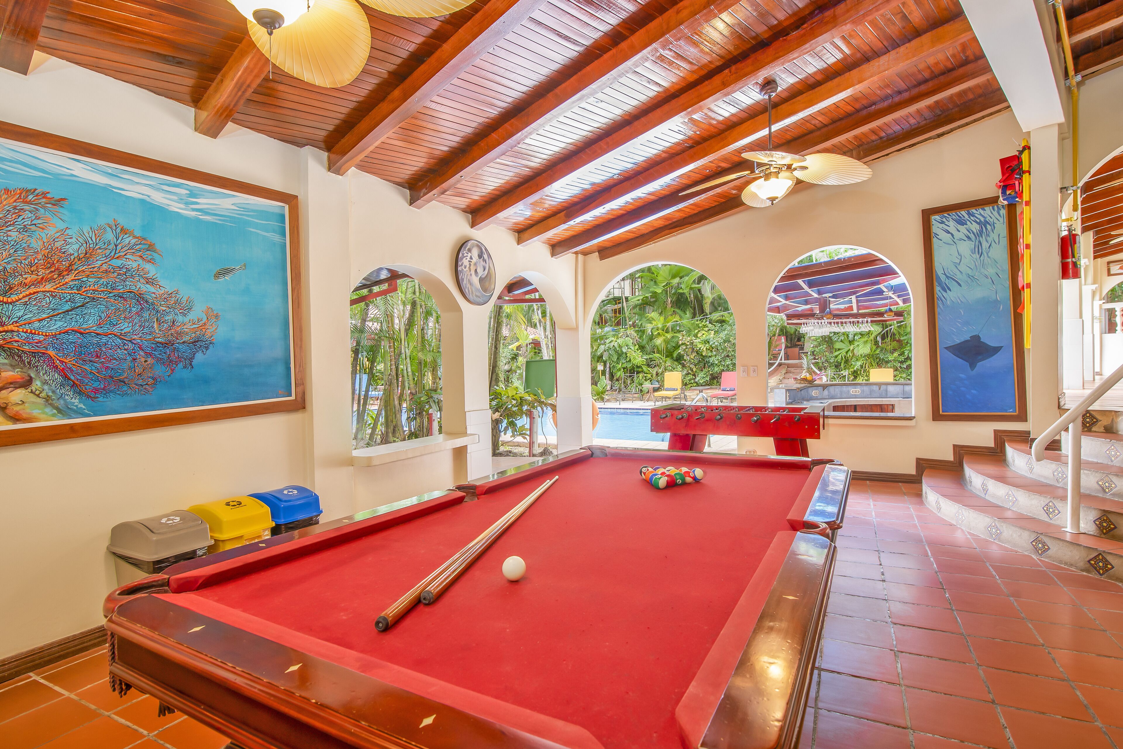 A pool table with a red felt surface, wooden ceiling beams, and a view of a pool area through an arched window.
