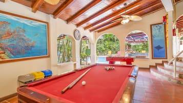 A pool table with a red felt surface, wooden ceiling beams, and a view of a pool area through an arched window.