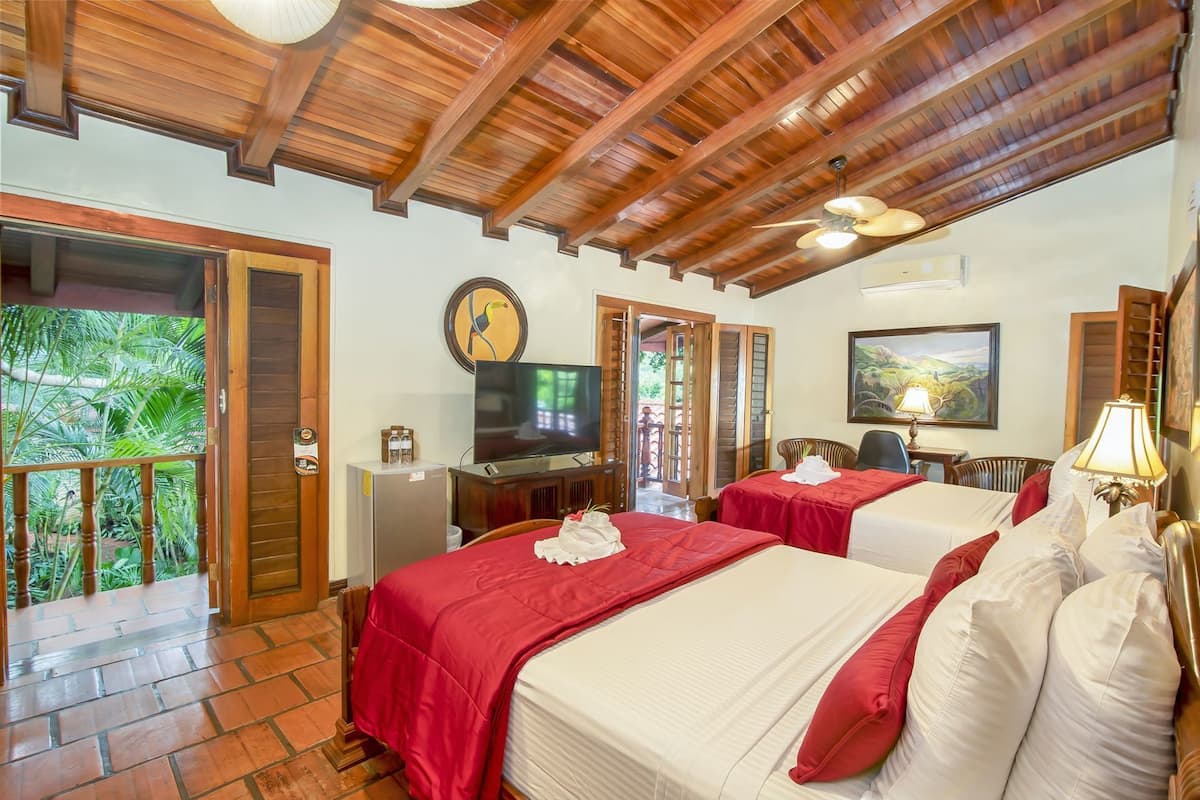 A hotel room with two beds, a television, a desk, and a balcony with a view of greenery.