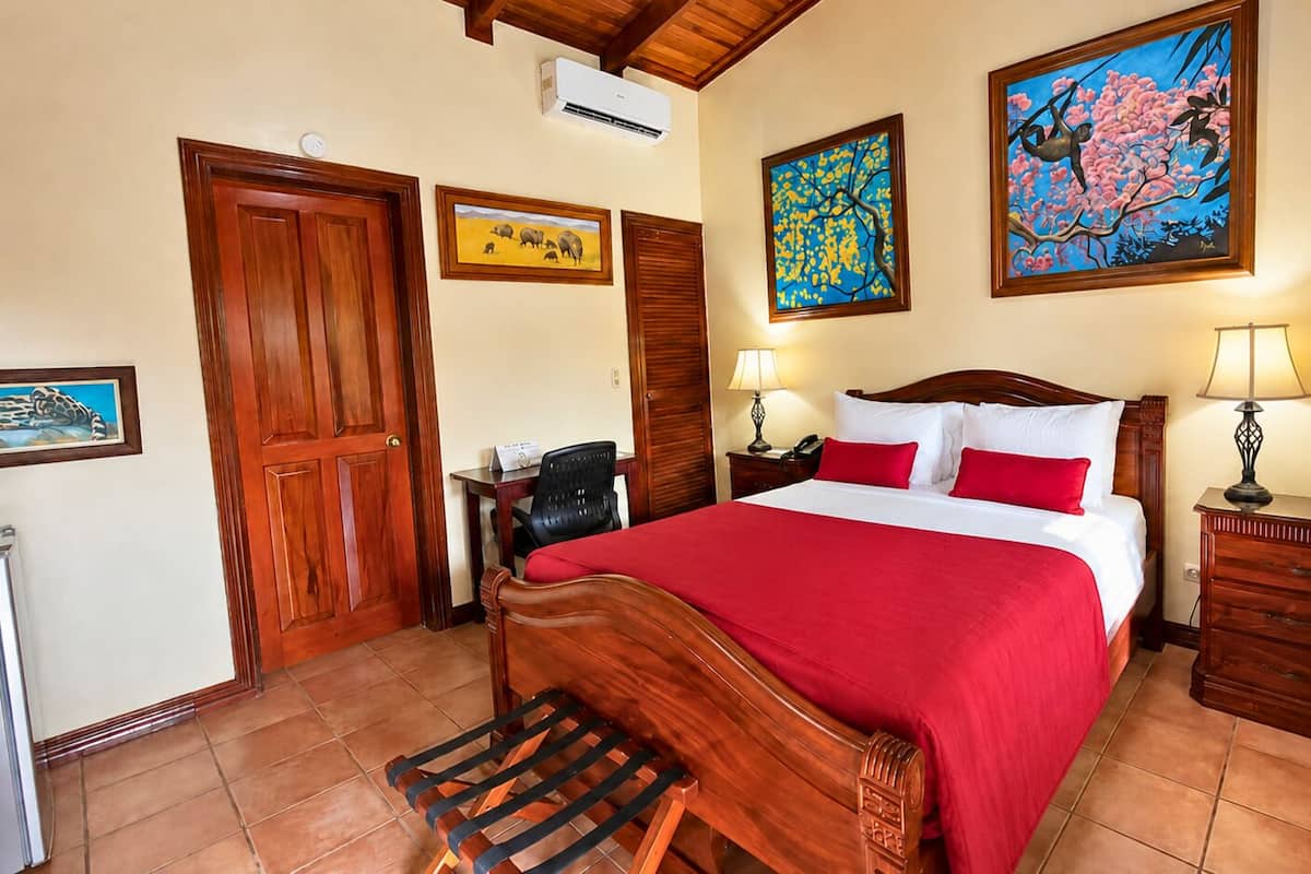 A hotel room with a wooden bed, red bedding, a desk with a chair, a dresser, and artwork on the walls.