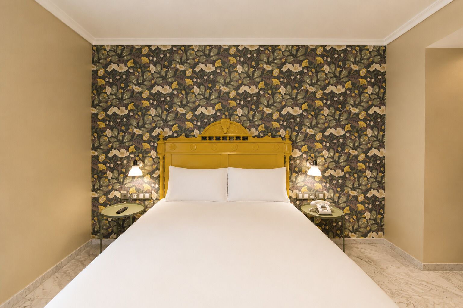 Une chambre d’hôtel avec un grand lit, un papier peint à motifs floraux, deux tables de chevet avec des lampes et un sol en marbre.