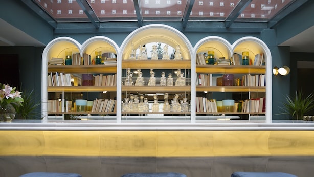 A bar area with a central counter, three blue bar stools, and a bookshelf filled with books and decorative items. - Petit Palace Puerta de Triana (Seville)