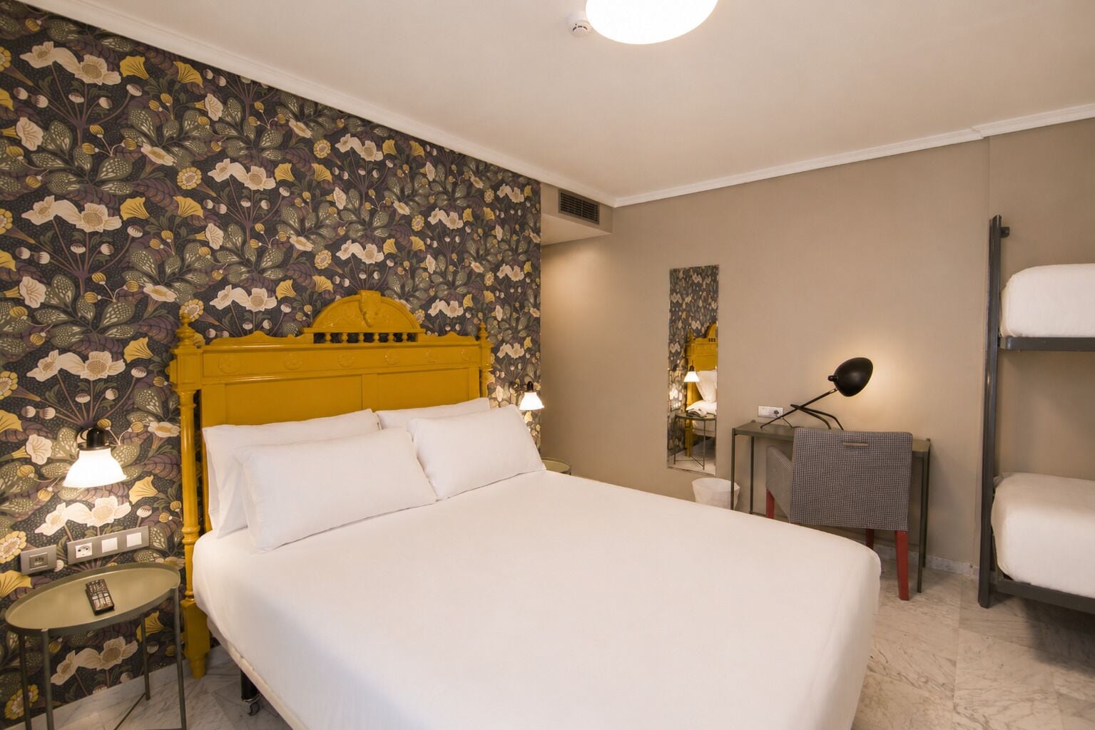 A hotel room with a large bed, a bedside table with a lamp, a mirror, a small table with a remote control, and a bunk bed.