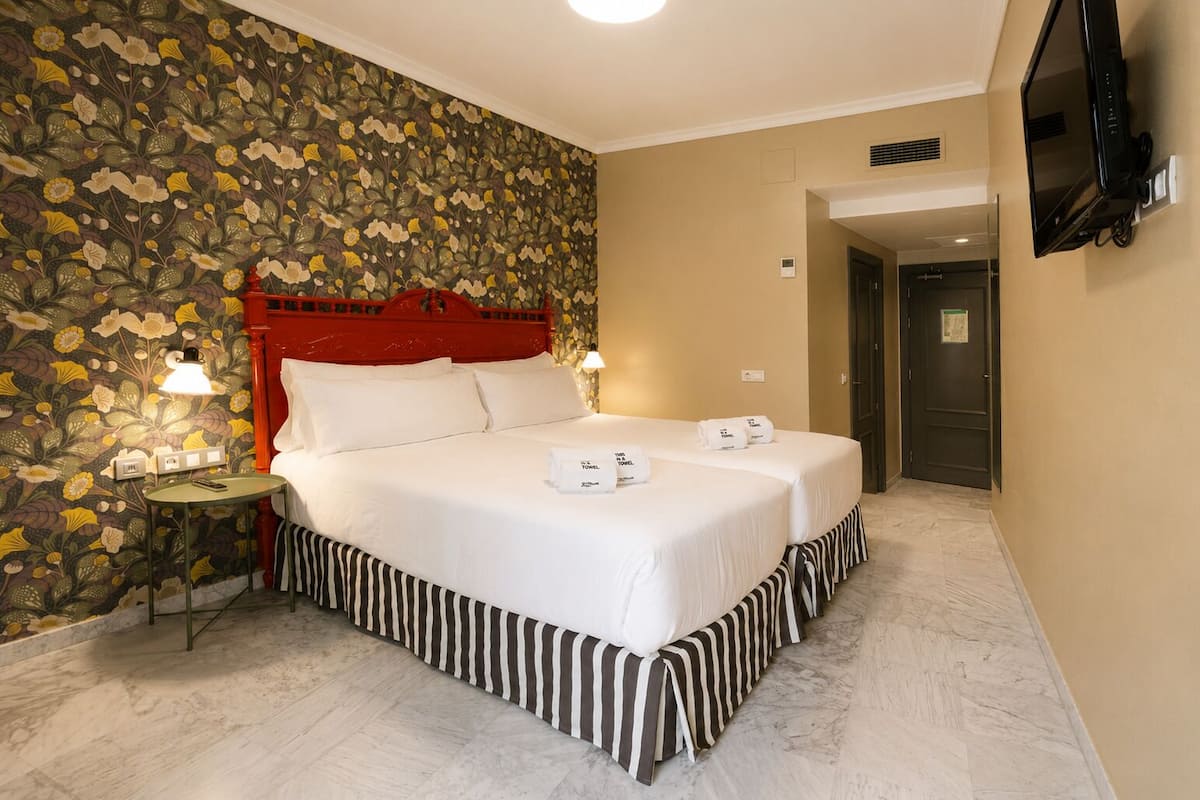 Une chambre d’hôtel avec un lit, deux tables de chevet, une tête de lit rouge, un papier peint à motifs floraux, une télévision et une porte.