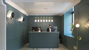 A reception desk with two staff members, a modern light fixture above, and a window with a view. - Petit Palace Puerta de Triana (Seville)