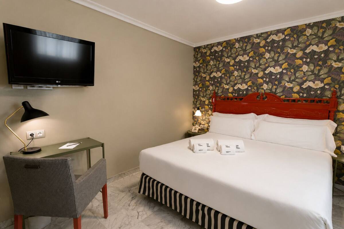 Une chambre d’hôtel avec un lit, un bureau doté d’une lampe, une télévision et un papier peint à motifs floraux.