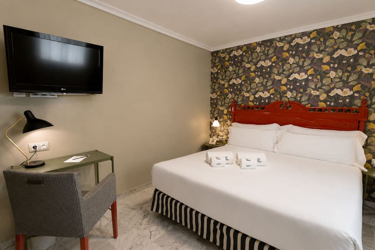 Une chambre d’hôtel avec un lit, un bureau doté d’une lampe, une télévision et un papier peint à motifs floraux.