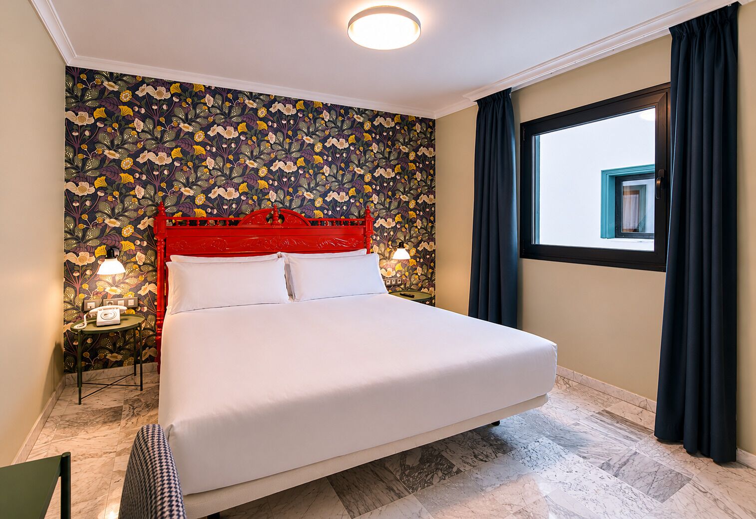 A hotel room with a bed, floral wallpaper, a red headboard, two bedside lamps, a window with curtains, and a marble floor.