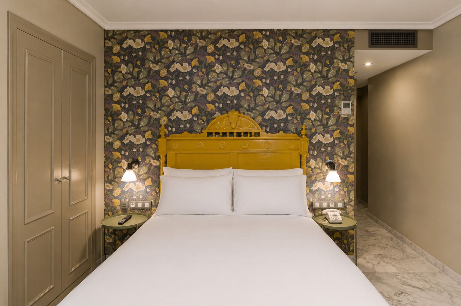 A hotel room with a large bed, floral wallpaper, a yellow headboard, two bedside lamps, and a marble floor.