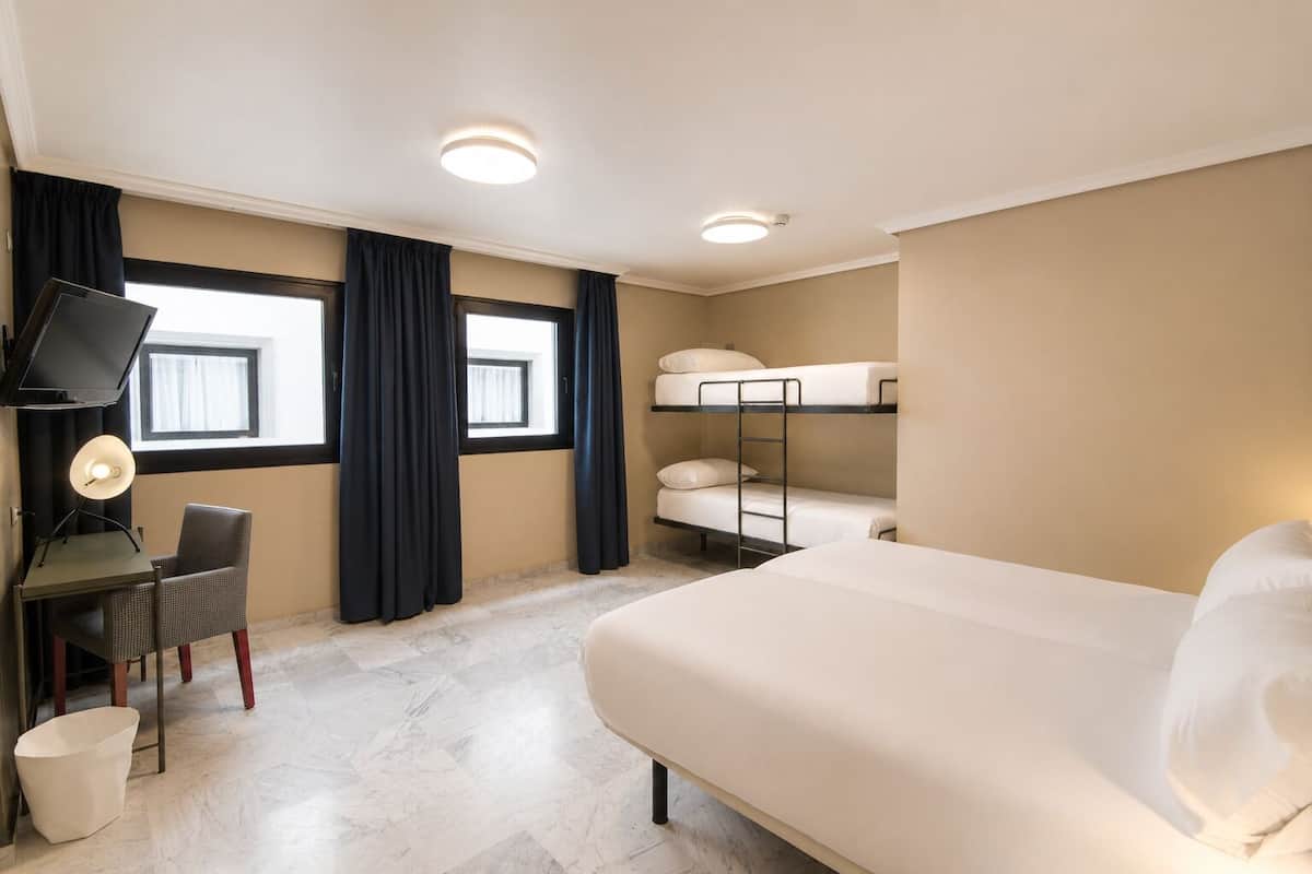 Une chambre d’hôtel comprenant un lit, des lits superposés, un bureau avec une chaise, une télévision et une fenêtre avec des rideaux.