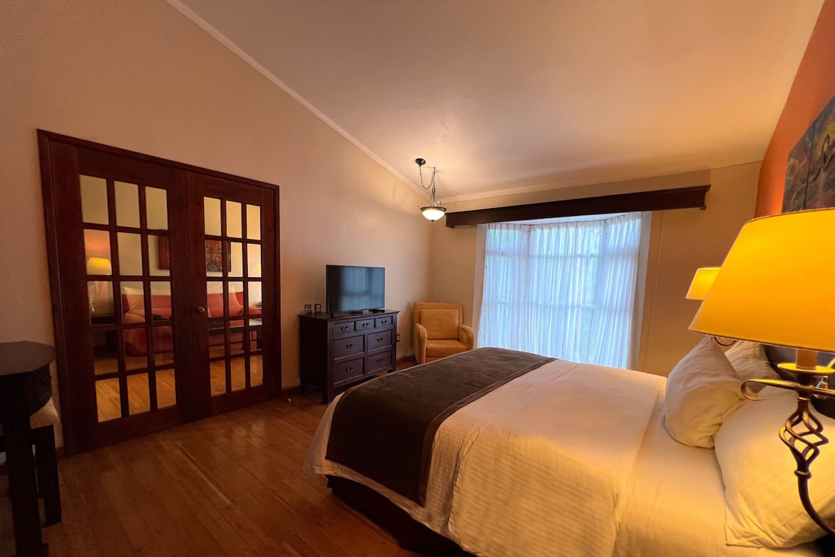 Une chambre Ă coucher avec un lit, une commode, un tĂ©lĂ©viseur, une chaise et une fenĂȘtre avec des rideaux.