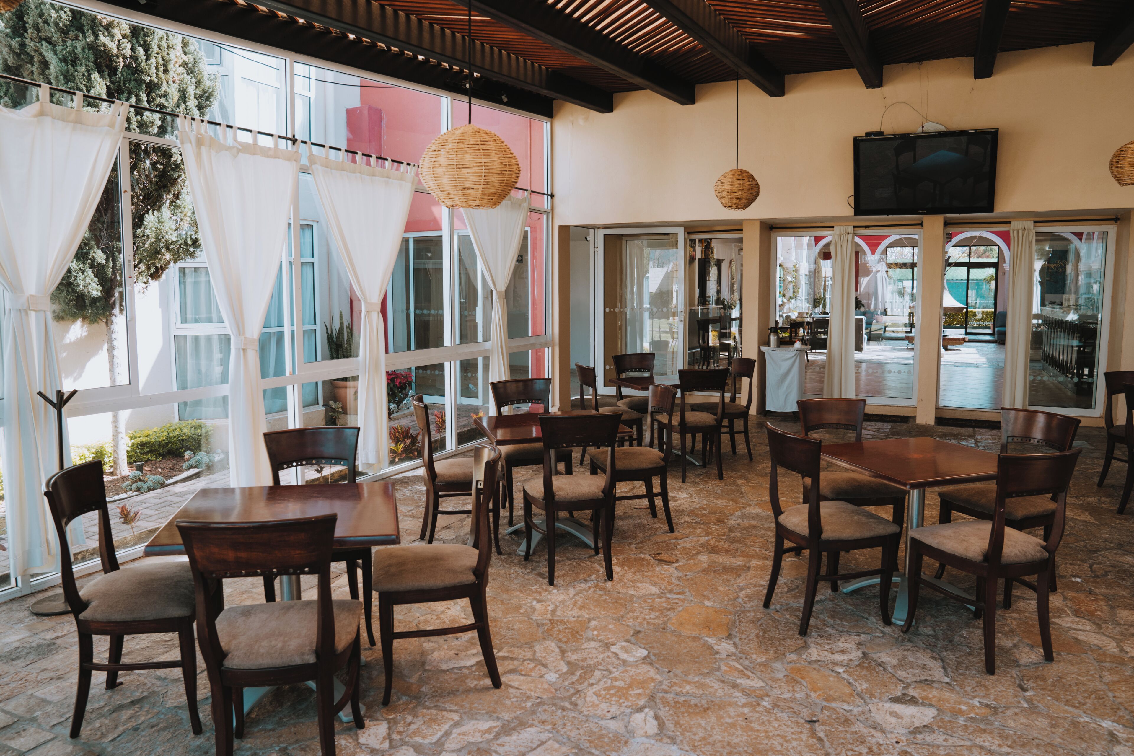 Un restaurante con suelo de piedra, mesas y sillas de madera, ventanas amplias con cortinas blancas y lámparas de mimbre colgantes.