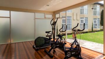 Un gimnasio con dos bicicletas estáticas y una bicicleta fija, ventanas amplias con vistas a un edificio y un césped.