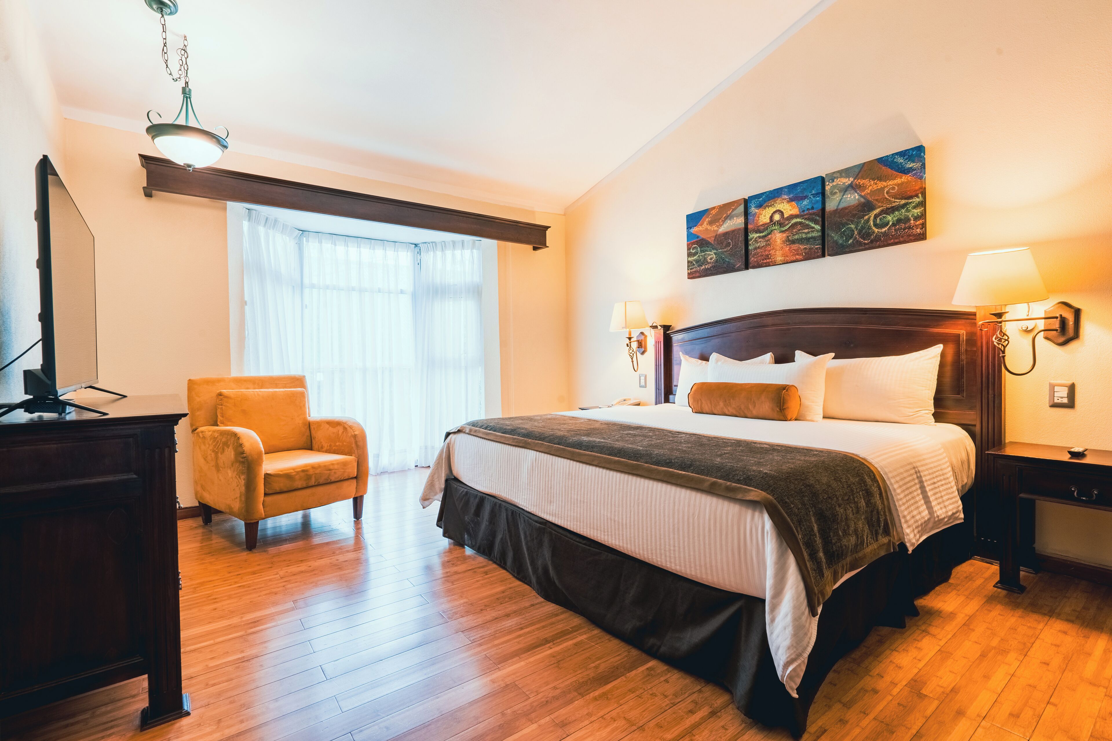 Una habitación de hotel con una cama grande, un escritorio de madera, una silla y un ventanal con cortinas.