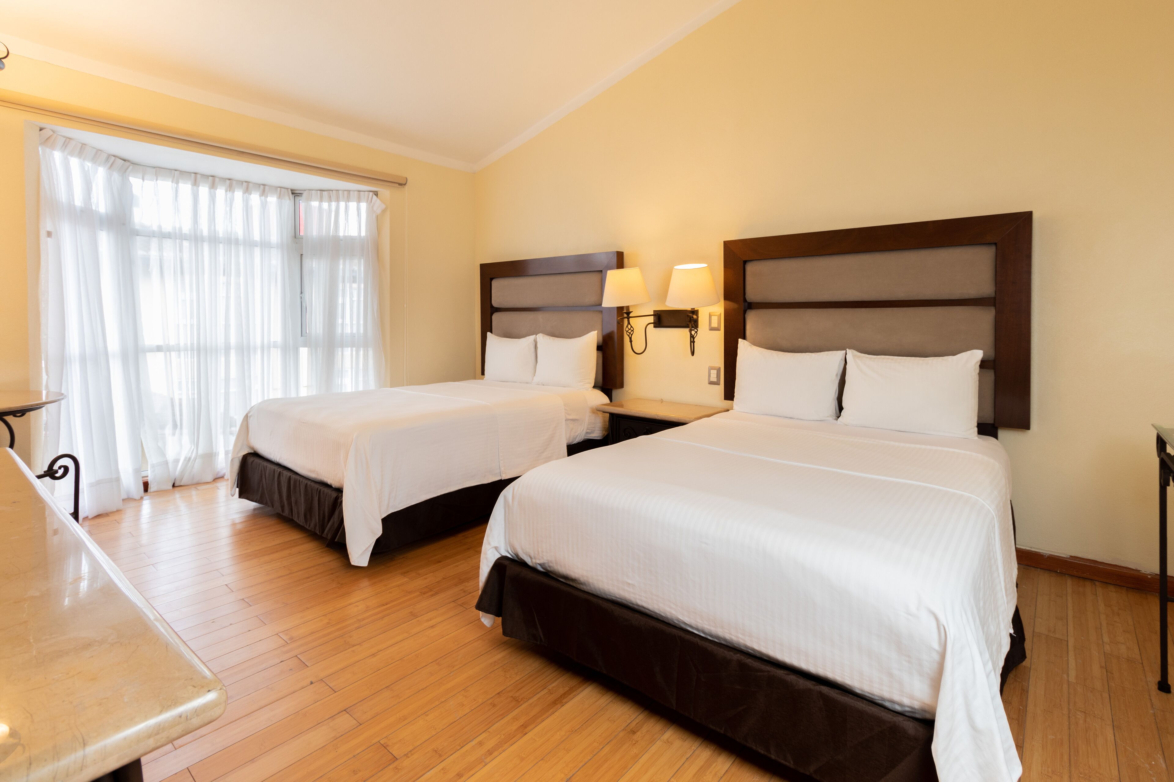 Una habitación de hotel con dos camas, suelo de madera, escritorio y ventana con cortinas traslúcidas.