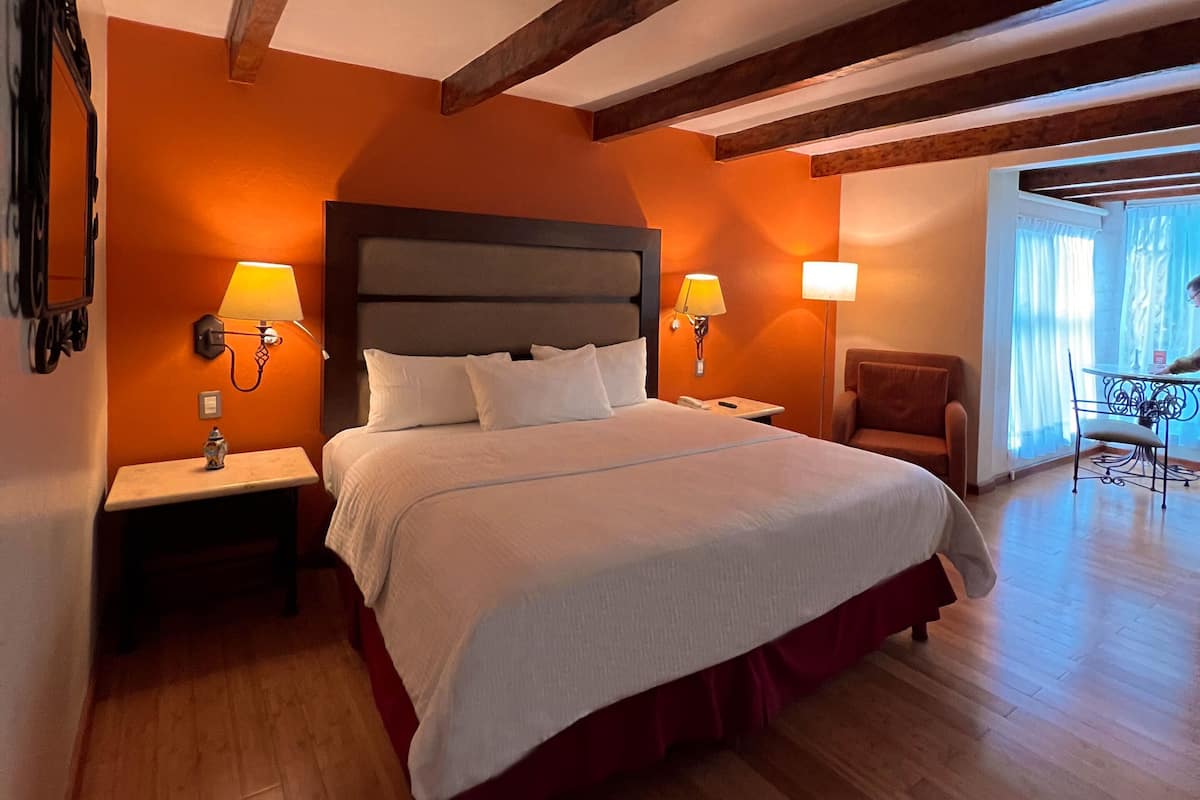Une chambre dâhĂŽtel avec un grand lit, des poutres en bois, deux lampes de chevet, une chaise et une vue sur lâextĂ©rieur.