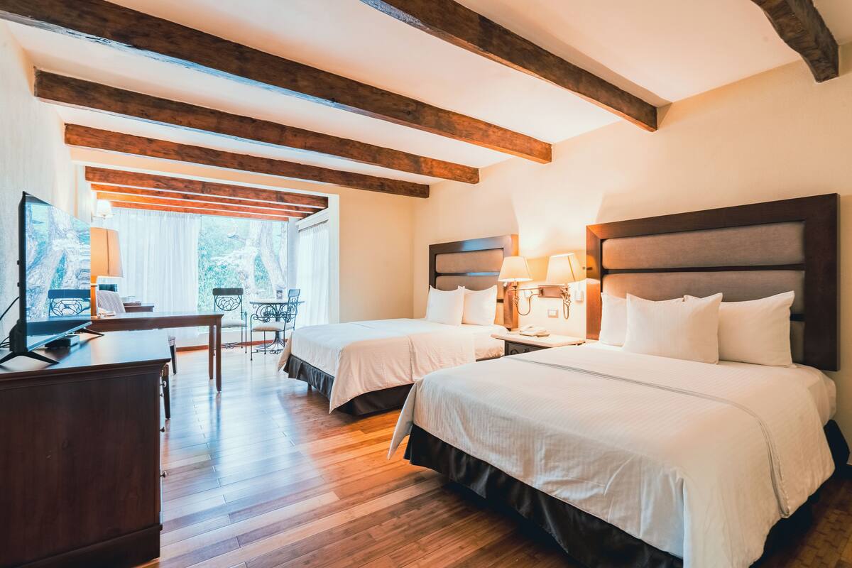 Camera d'albergo con due letti, travi in legno al soffitto, una scrivania con un computer e una finestra con tende.