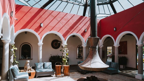 A spacious atrium with a high ceiling, red walls, and white arches. There are seating areas with sofas and chairs, and a large decorative fountain in the center.