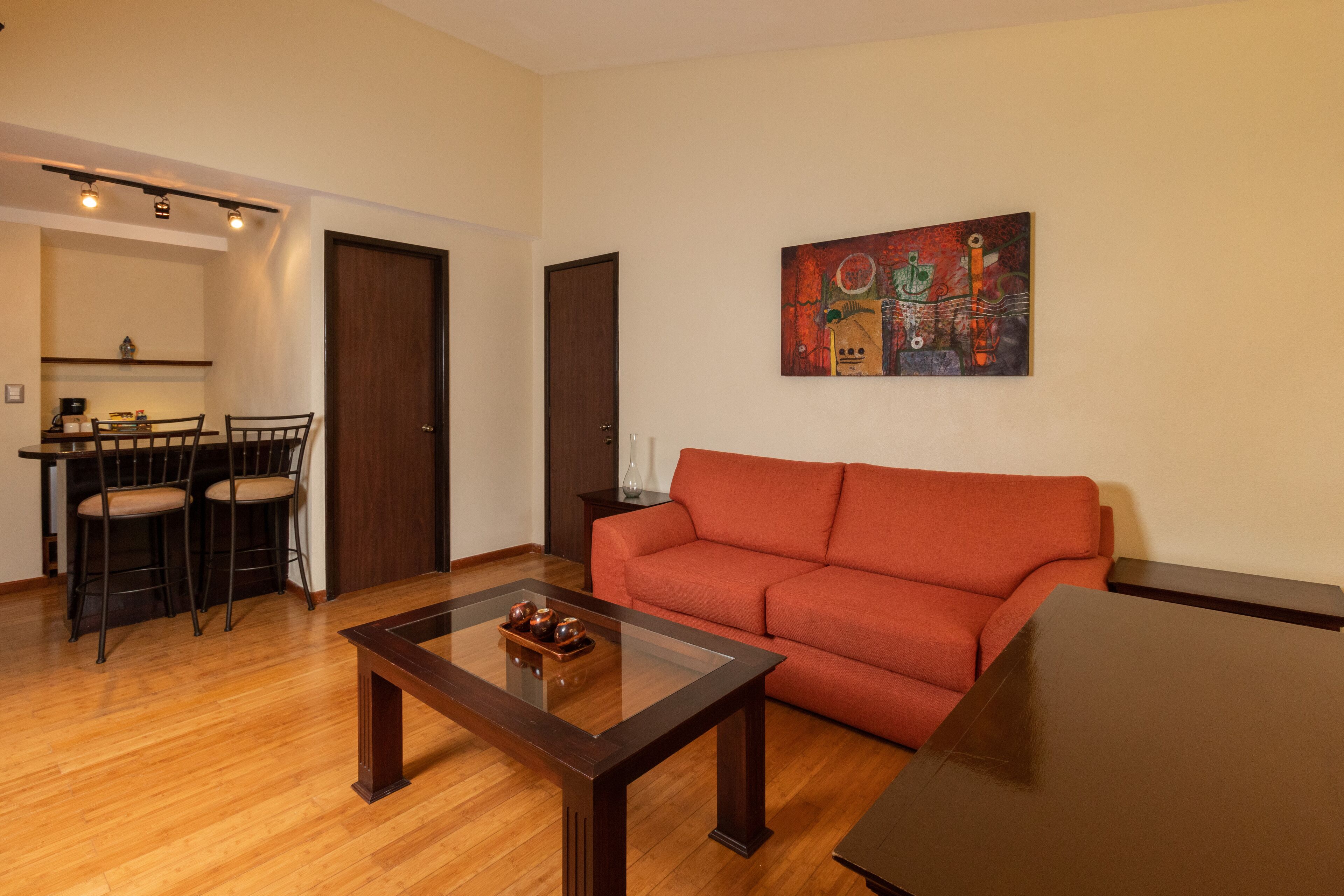 Un salon avec un plancher en bois, un canapé rouge, une table basse en verre et un tableau accroché au mur. Au fond, on aperçoit un coin repas avec une table et des chaises.