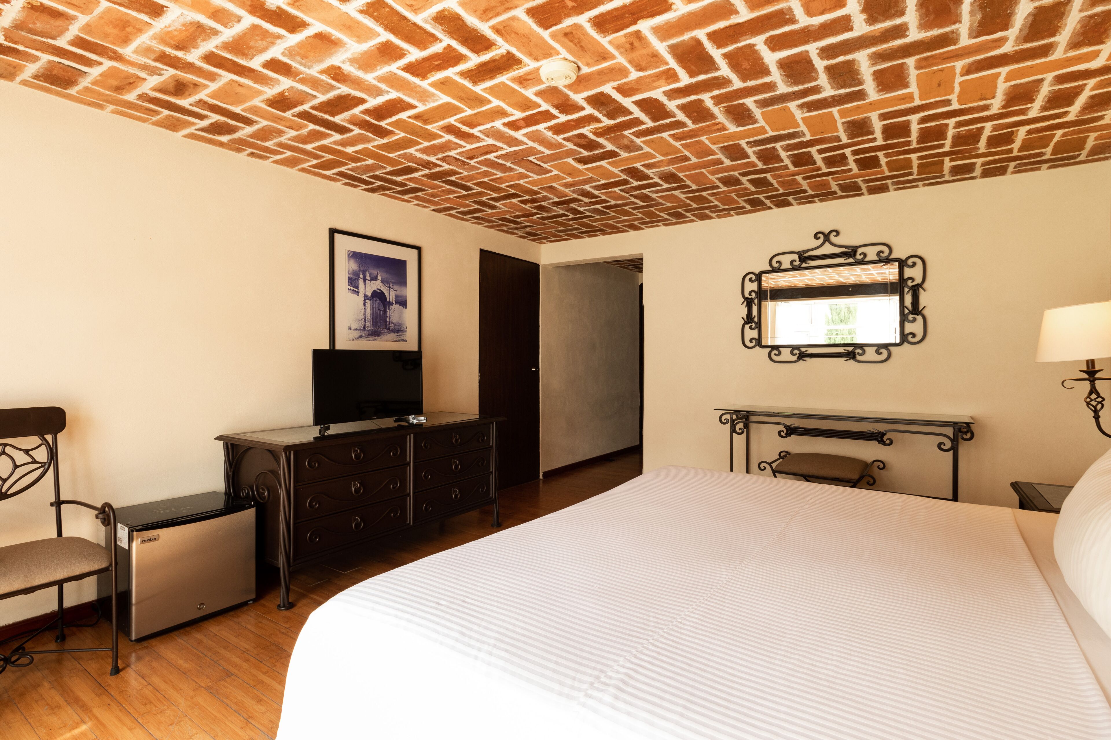 Habitación de hotel con techo de ladrillo, una cama, un aparador, un televisor y un cuadro enmarcado en la pared.