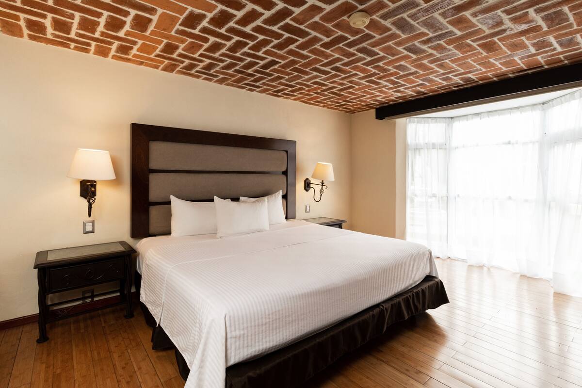Camera d'albergo con soffitto in mattoni, un letto grande con lenzuola bianche, due comodini con lampade e un'ampia finestra con tende leggere.