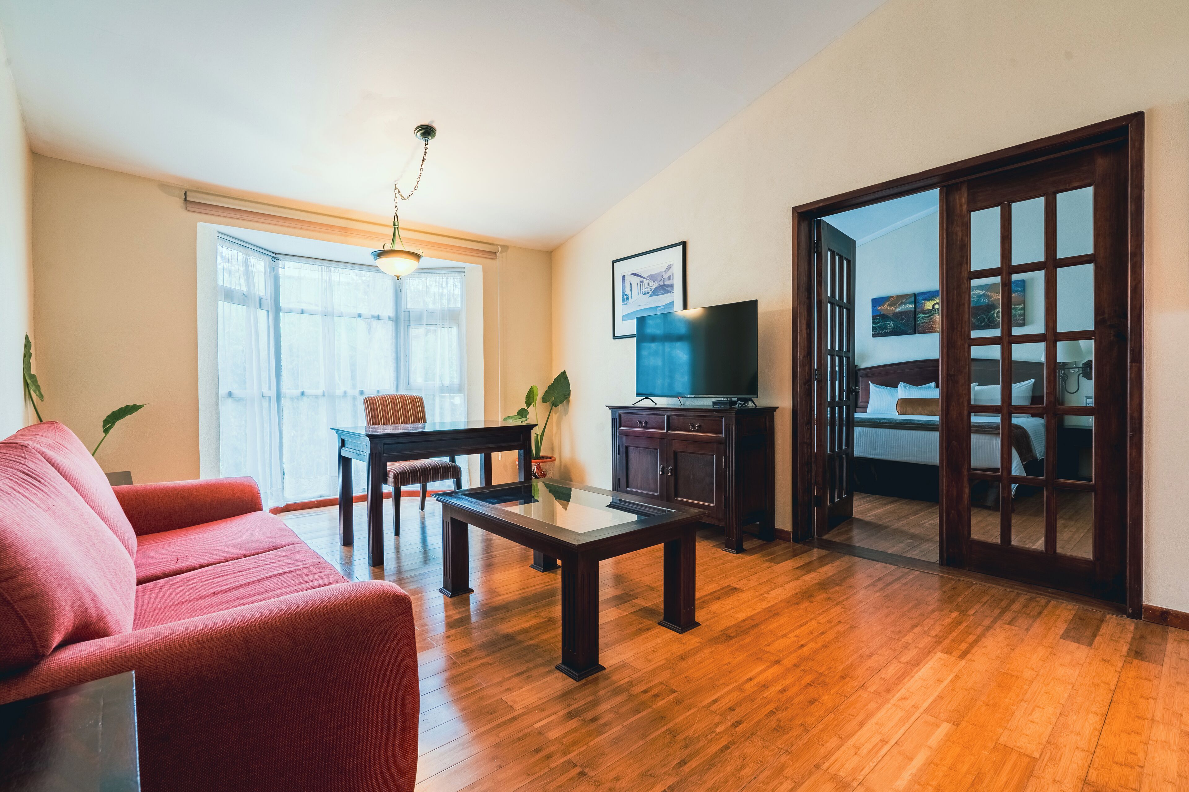 A living room with a red sofa, a glass-top coffee table, a wooden floor, a TV, and a sliding door leading to a bedroom.