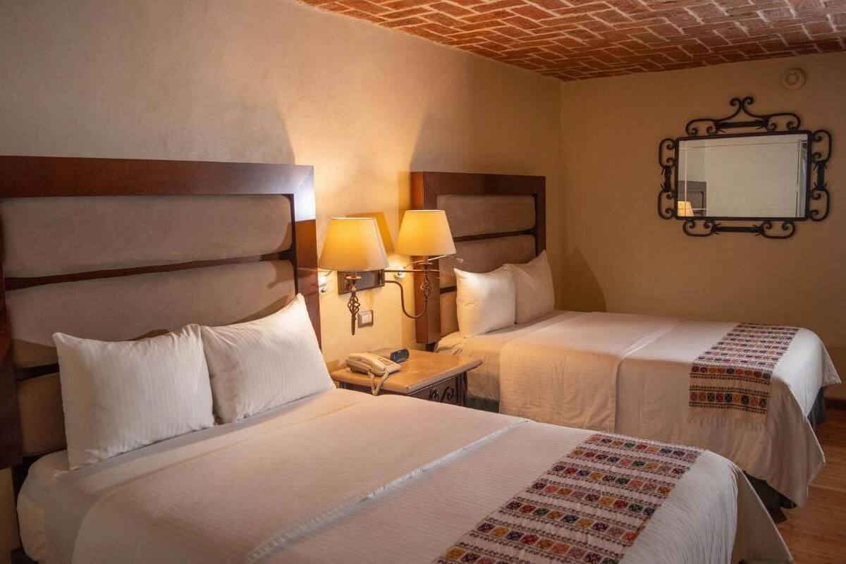Camera d'albergo con due letti, ognuno con lenzuola bianche e una coperta decorata. Sono presenti lampade sui comodini e un piccolo tavolo con un telefono.