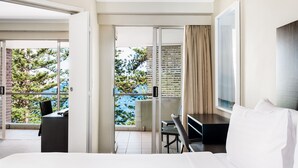 Habitación de hotel con una cama, un escritorio y una silla. Tiene un balcón con vistas a árboles y un edificio.