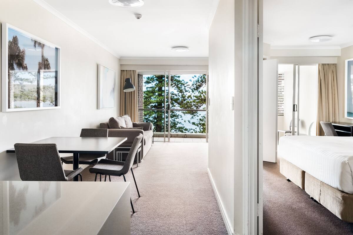 A hotel room with a large bed, a dining area with a table and chairs, a sofa, and a view of trees through the window.