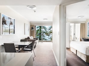 A hotel room with a large bed, a dining area with a table and chairs, a sofa, and a view of trees through the window.
