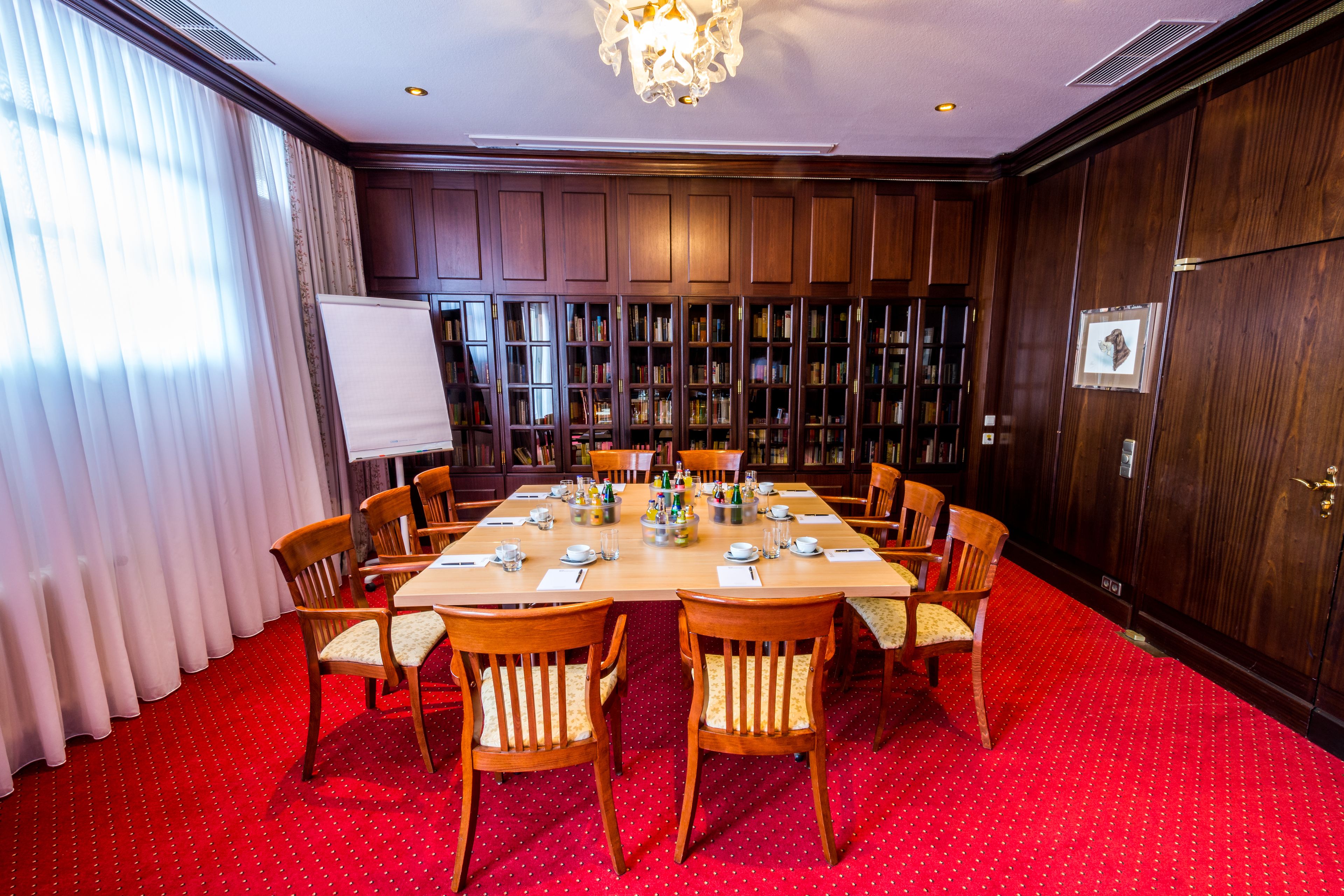 Une salle de conférence avec un tapis rouge, des boiseries, un lustre et un tableau blanc.