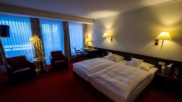 Quarto de hotel com uma cama grande, duas luminárias de cabeceira, uma escrivaninha com cadeira e uma TV fixada na parede.