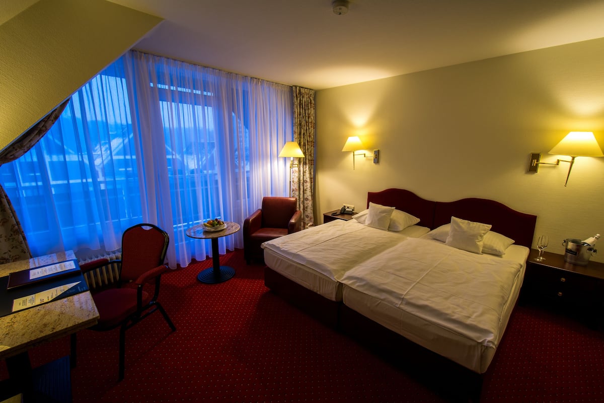 Ein Hotelzimmer mit einem großen Bett, zwei Nachttischlampen, einem Schreibtisch mit Stuhl, einem kleinen Tisch mit Lampe und Blick durch einen leichten Vorhang.
