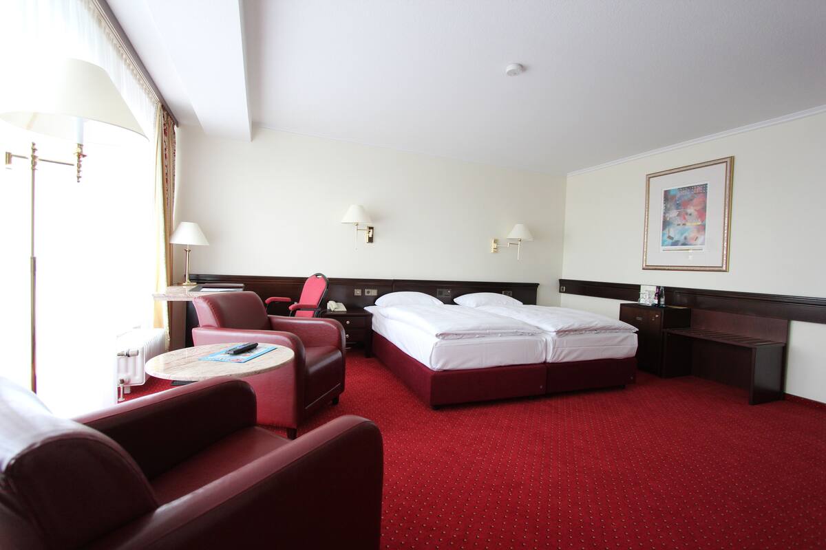 Ein Hotelzimmer mit rotem Teppich, einem Bett mit weißer Bettwäsche, einem Schreibtisch mit Lampe, einem Sessel und einem kleinen Tisch.