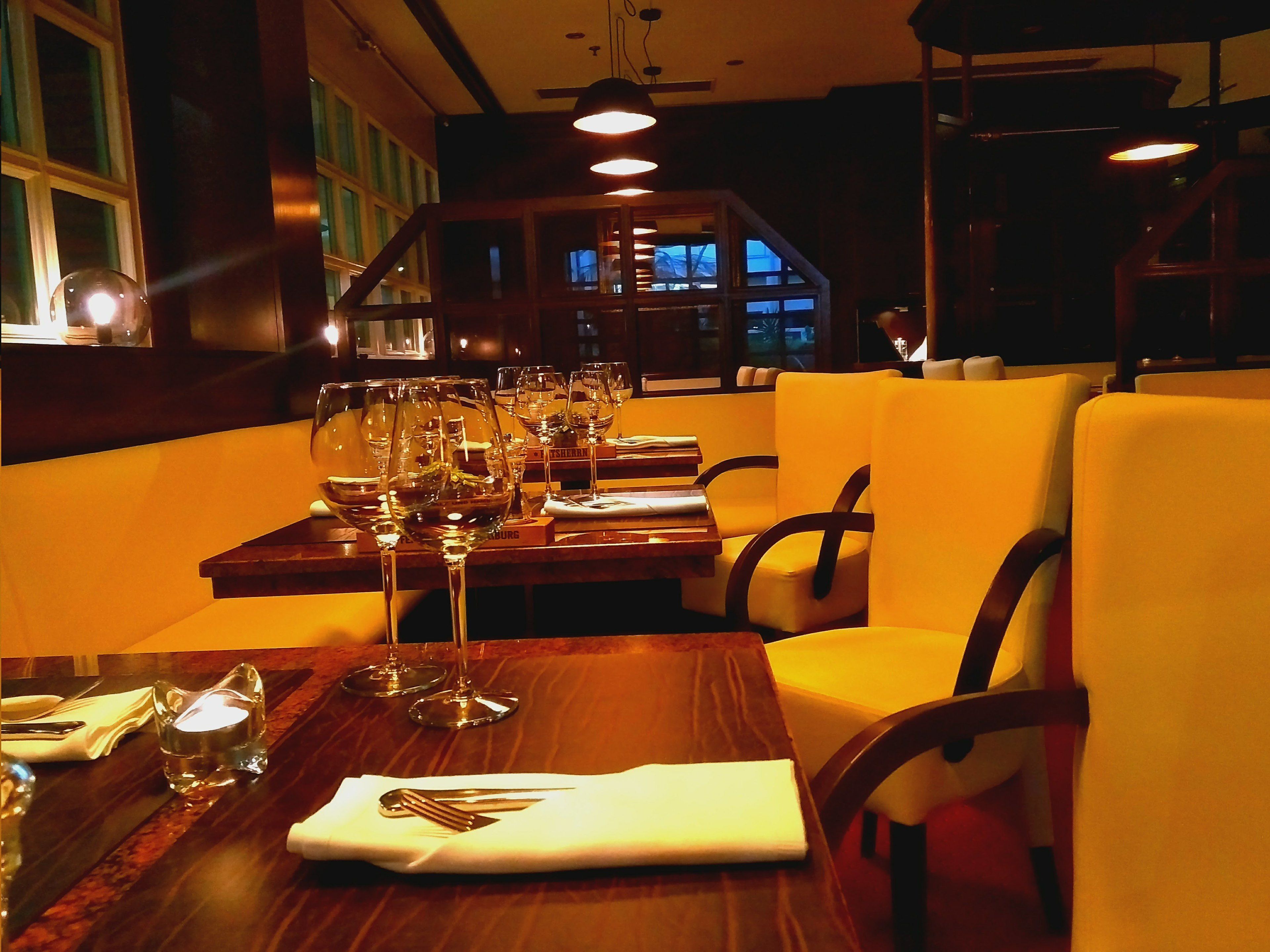 A restaurant interior with wooden tables, yellow upholstered chairs, and multiple wine glasses on each table.