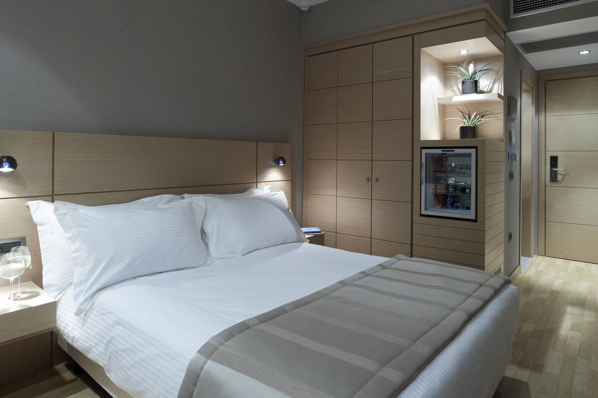 Una cama bien hecha con ropa blanca, una mesita de noche con un vaso de agua y un minibar empotrado con refrigerador y estantes.