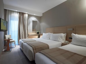 A hotel room with two beds, a desk, a chair, a TV, and a large window with curtains. - Anatolia Hotel (Komotini)