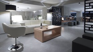 A modern living room with a white sofa, a wooden coffee table, and a black floor lamp. There is a fireplace with a fire, and a dining area with chairs and a table in the background. - Anatolia Hotel (Komotini)