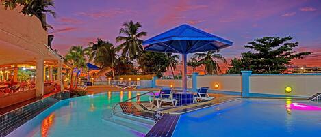 A pool area with lounge chairs and umbrellas, surrounded by palm trees and a sunset sky.