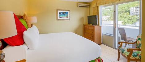Habitación de hotel con cama, televisor, sillón y balcón con vistas a la vegetación.