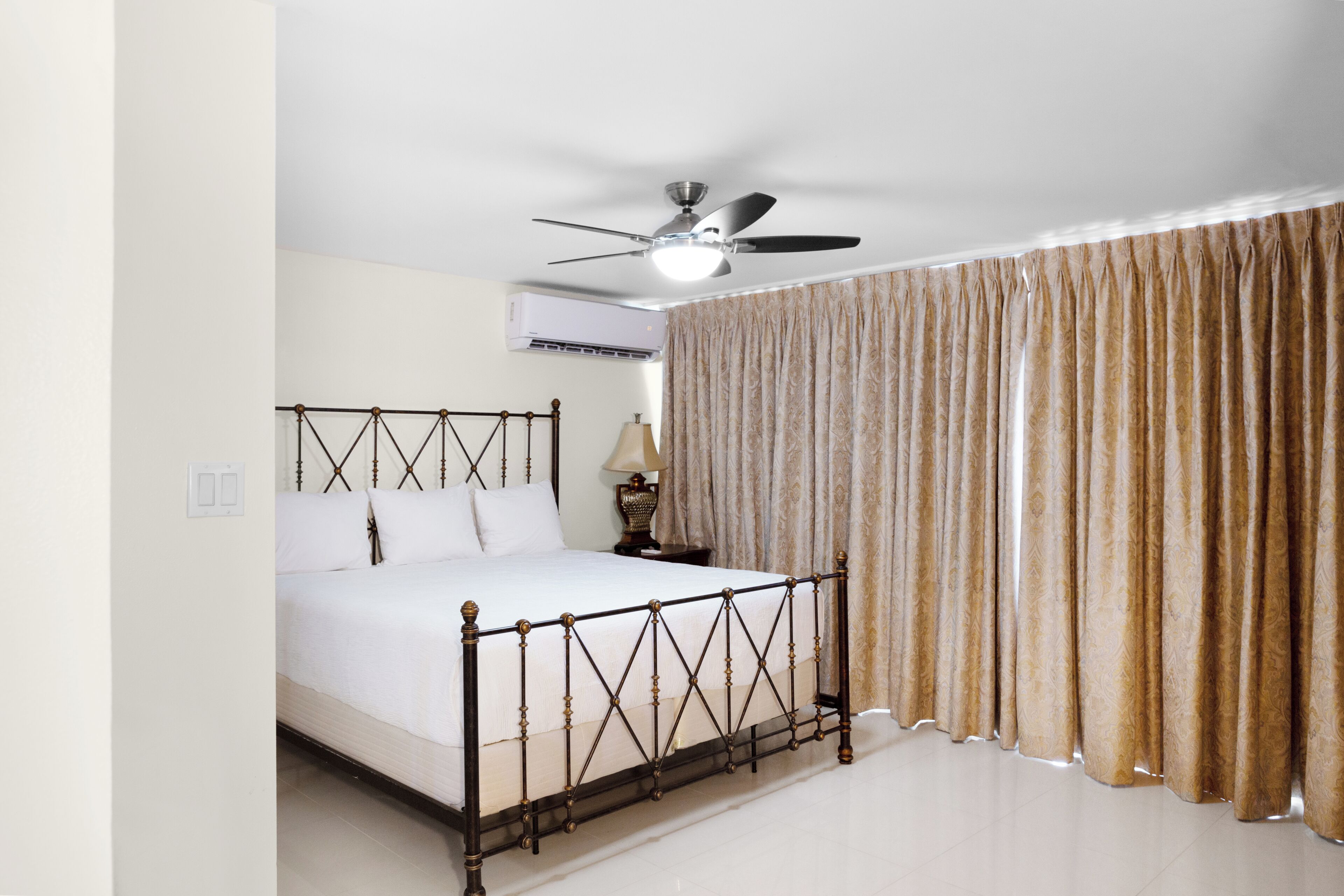 A neatly made bed with a headboard, a ceiling fan, an air conditioning unit, and a curtain.