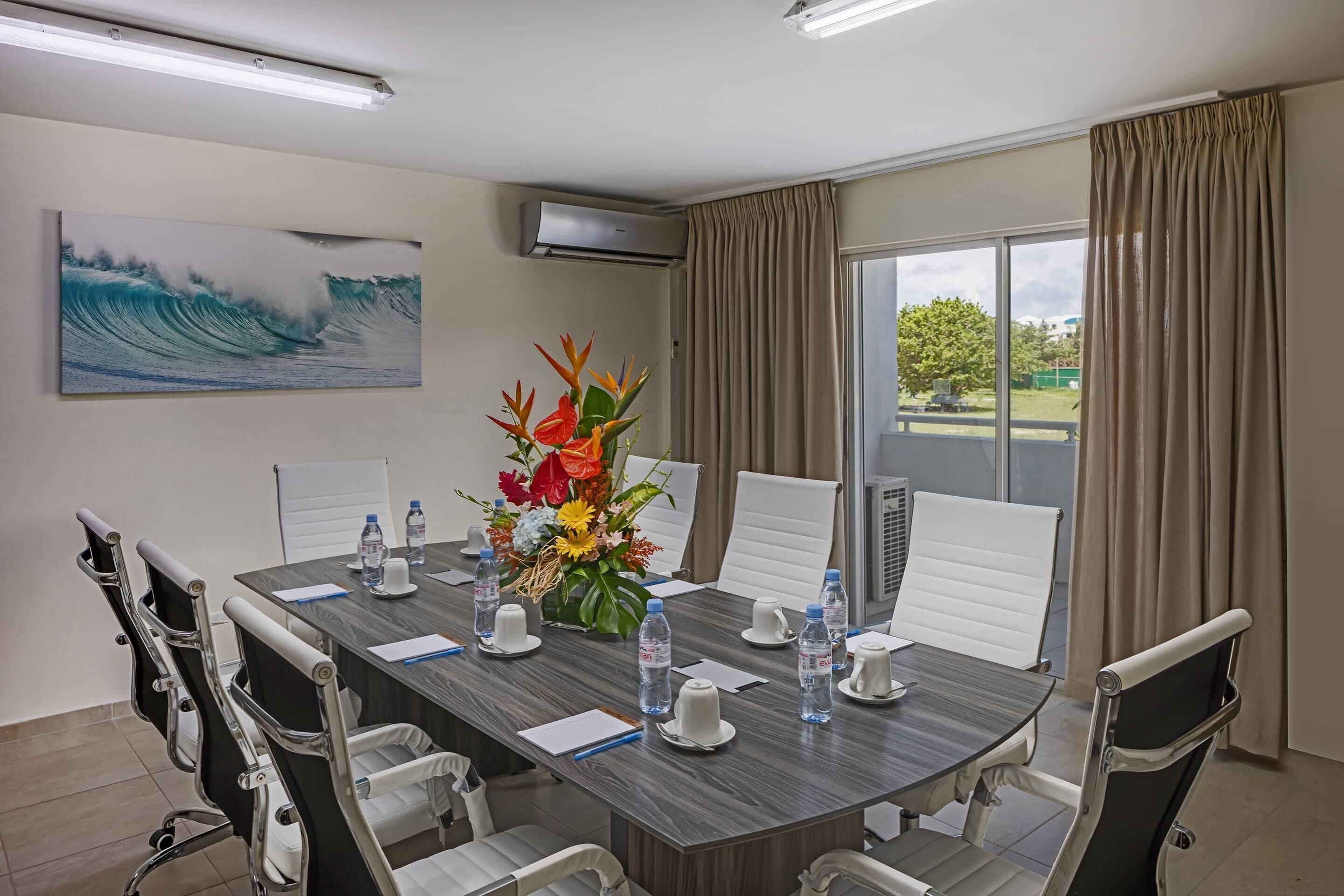 Uma sala de conferências com uma mesa comprida, cadeiras brancas, um arranjo floral, garrafas de água e uma grande pintura de ondas na parede.