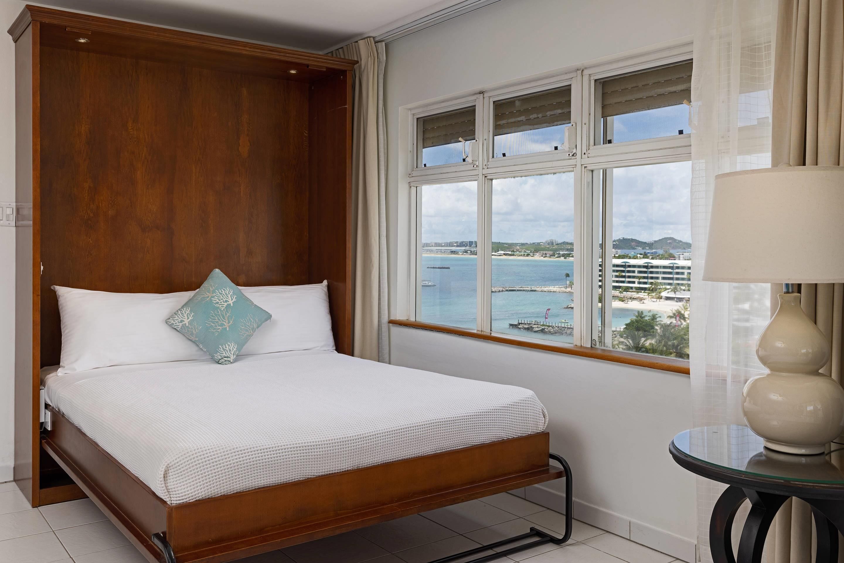 Quarto de hotel com cama de madeira, vista para o mar e uma mesinha com abajur.