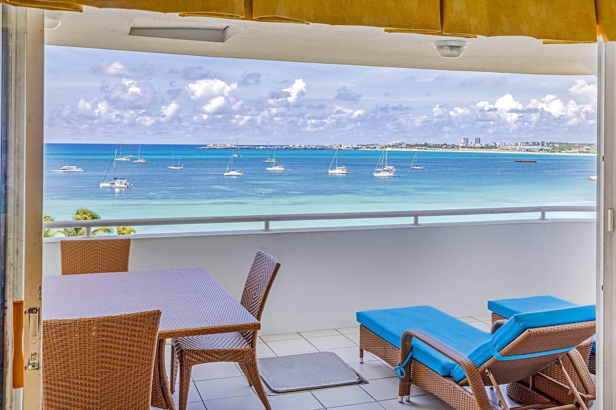 Un balcón con mobiliario de mimbre, vista al mar con barcos y un horizonte urbano distante.