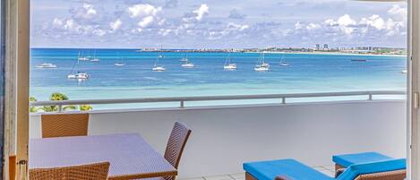 Un balcon agrémenté de meubles en osier, une vue sur la mer avec des bateaux, et un panorama sur la ville au loin.