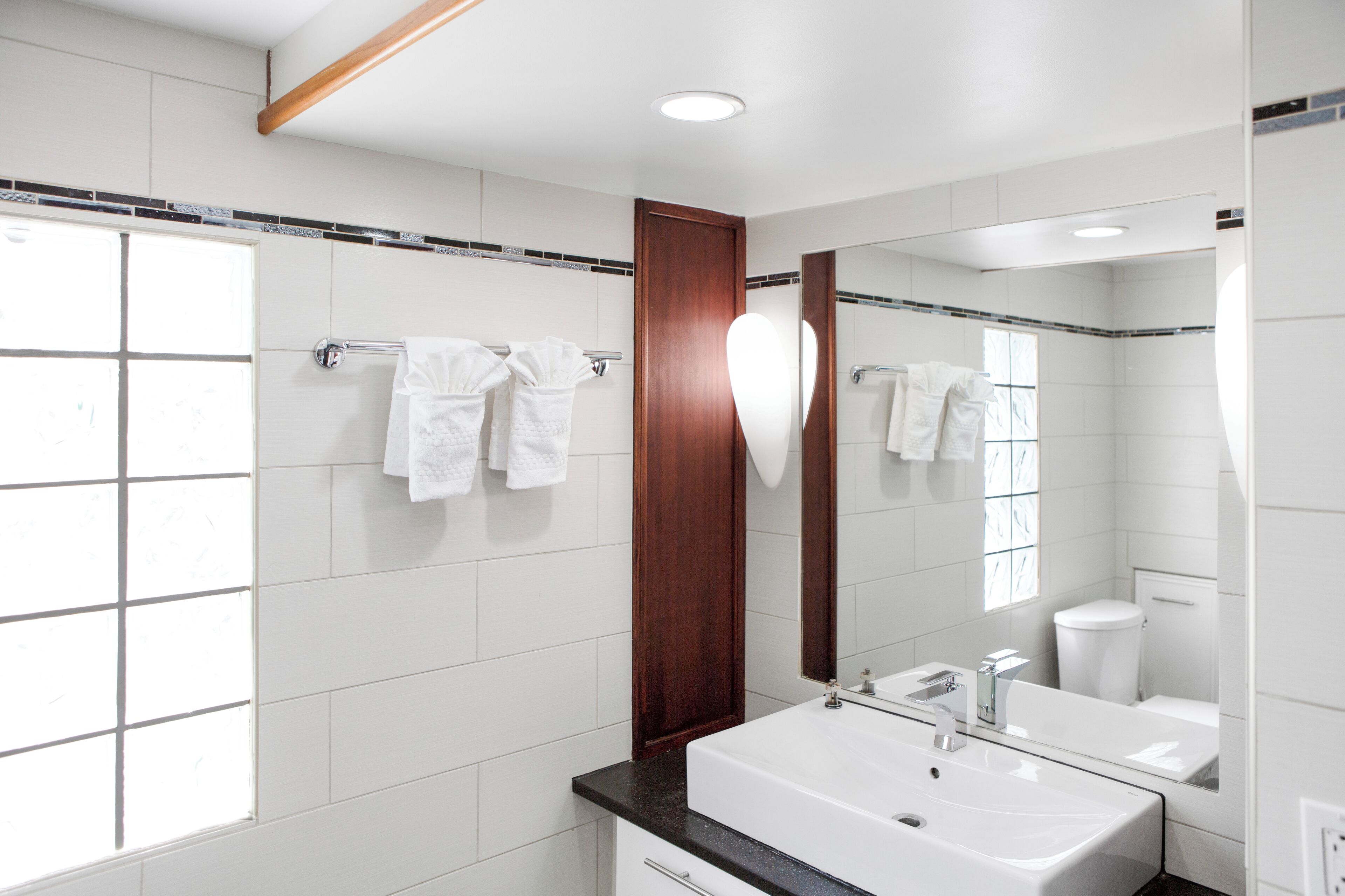 A modern bathroom with a large mirror, white tiles, a wooden door, and a black countertop.